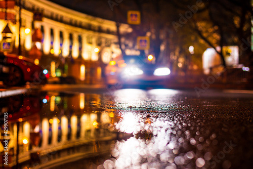 Autumn rainy night in the city. Headlights of a riding car. Parked cars. Residential buildings in the city center. Colorful colors. Close up view from the level of the puddle on the pavement.