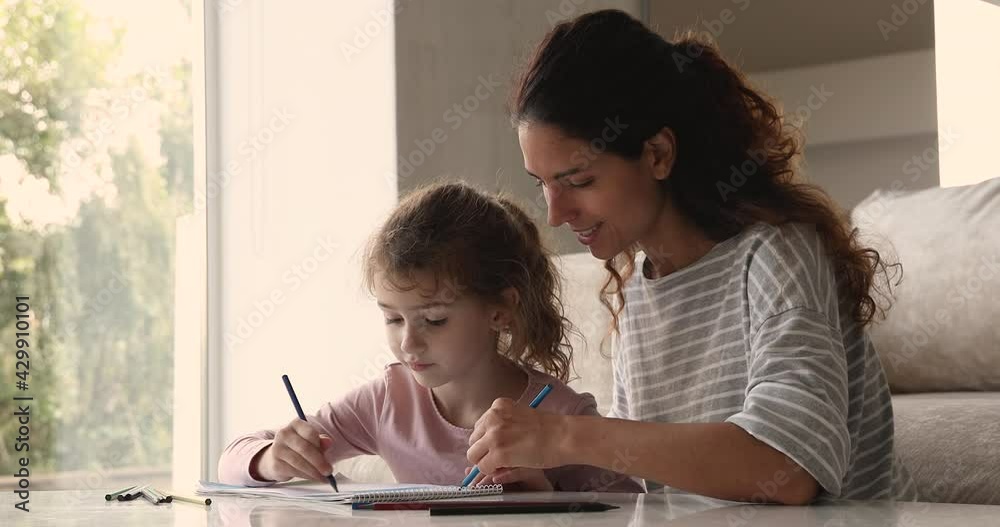 Caring young mom drawing with pencils teaching coloring talk to little daughter sit at table in living room, friendly nanny helping child create painting. Children development, creative hobby concept Caring young mom drawing with pencils teaching coloring talk to little daughter sit at table in living room, friendly nanny helping child create painting. Children development, creative hobby concept