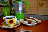 Zdjęcie przedstawiające filiżankę kawy z wafelkami na stole z kawiarką w tle