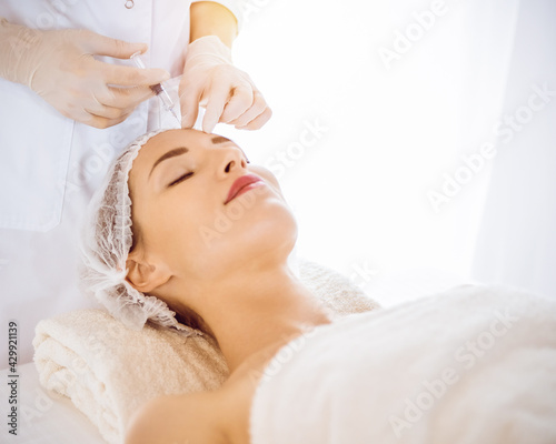 Beautiful woman receiving beauty injections with closed eyes in sunny medical center. Beautician doctor hands doing beauty procedure to female face with syringe. Cosmetic medicine and surgery concept