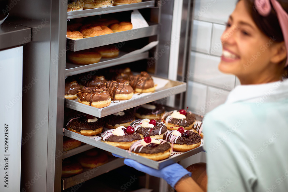 A young small business female owner in a candy workshop fills the closet with delicious handmade donuts with satisfaction. Pastry, dessert, sweet, making