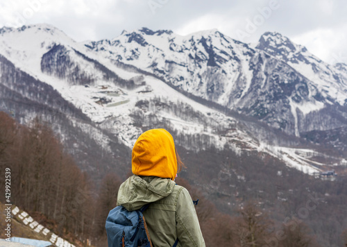 Young woman in yellow hood with backpack from behind enjoying the view of the Caucasian mountain peaks covered with snow, hiking, active lifestyle, landscape