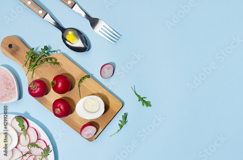 Recipe of organic salad of radish, arugula and eggs with olive oil on blue background, flat lay, copy space. Modern concept with shadow and hard light.