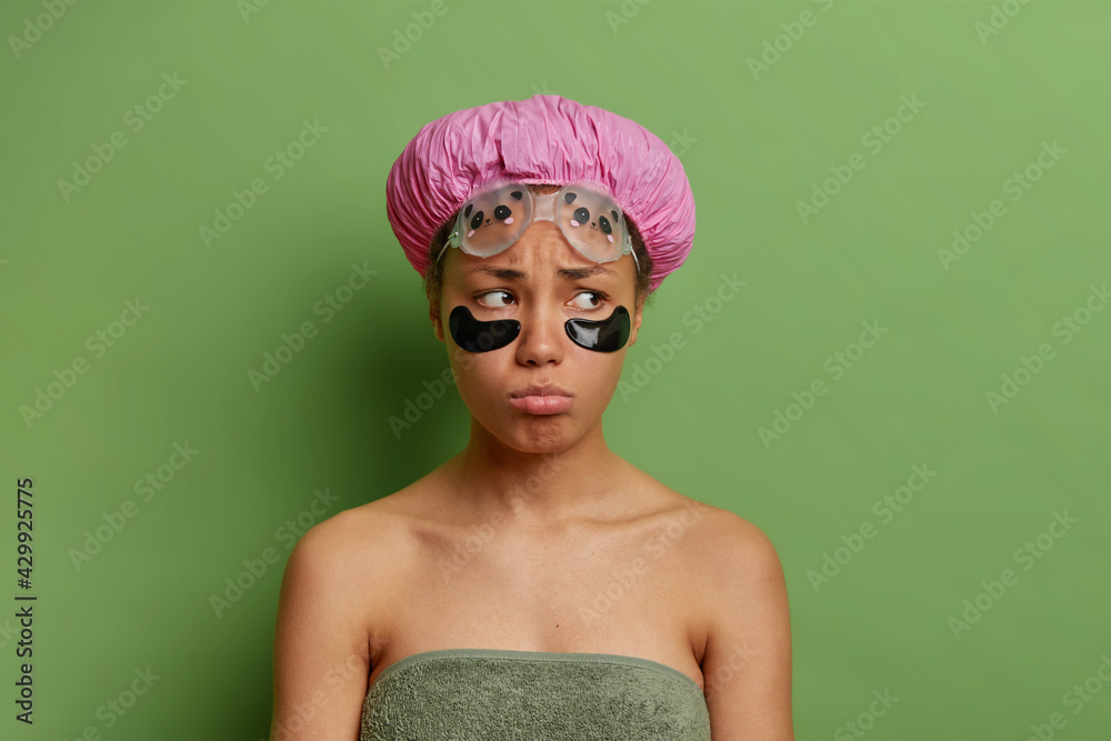 Studio shot of displeased ethnic woman looks with gloomy expression undergoes beauty procedures after taking shower wrapped in towel wears bath hat isolated over green background. Face care concept Studio shot of displeased ethnic woman looks with gloomy expression undergoes beauty procedures after taking shower wrapped in towel wears bath hat isolated over green background. Face care concept