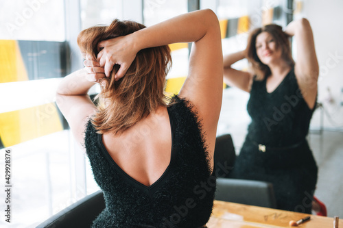 Beautiful smiling young woman plus size body positive in black evening dress make hair style near mirror at the beauty studio photo