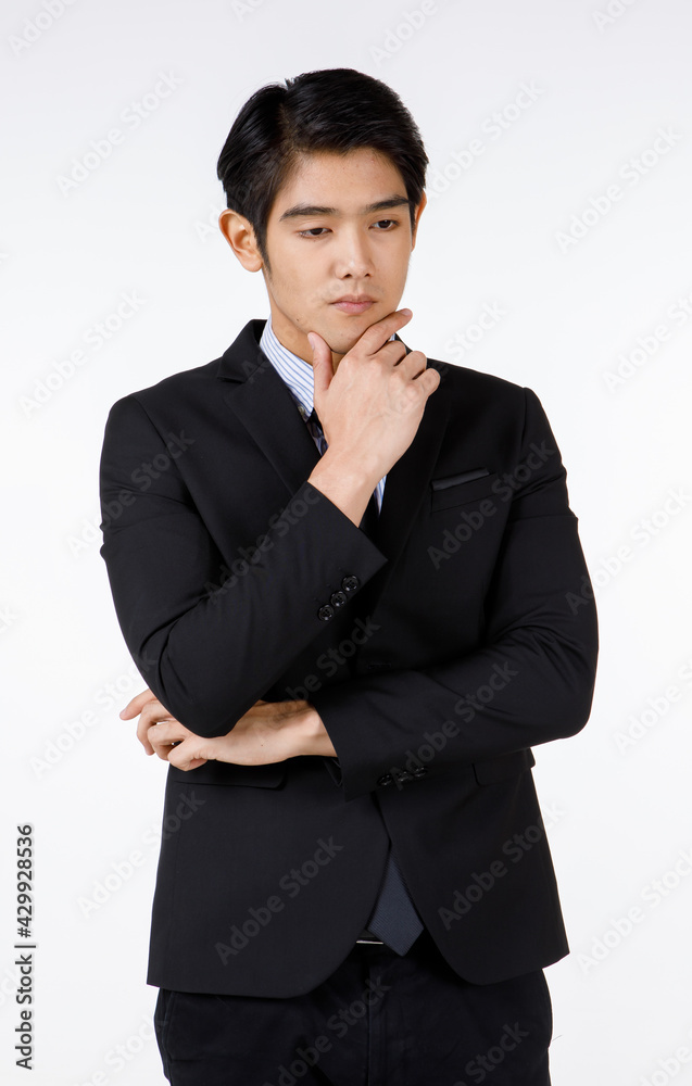 Portrait shot of Asian young handsome happy intelligence successful businessman in black formal suit stand hold hand touch chin brooding thinking alone in front of white wall background