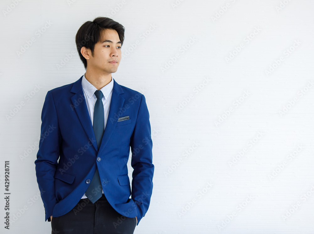 Portrait closeup shot of Asian young handsome happy intelligence college teen student wears blue formal suit and necktie standing hold hands in pants pockets look outside in front of white wall Portrait closeup shot of Asian young handsome happy intelligence college teen student wears blue formal suit and necktie standing hold hands in pants pockets look outside in front of white wall