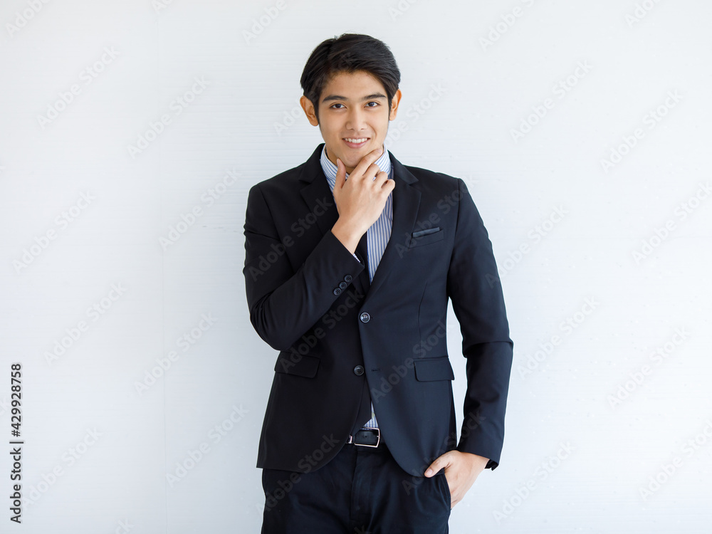 Portrait shot of Asian young handsome happy intelligence successful businessman in black formal suit stand hold hand touch chin brooding thinking and smiling alone in front of white wall background Portrait shot of Asian young handsome happy intelligence successful businessman in black formal suit stand hold hand touch chin brooding thinking and smiling alone in front of white wall background