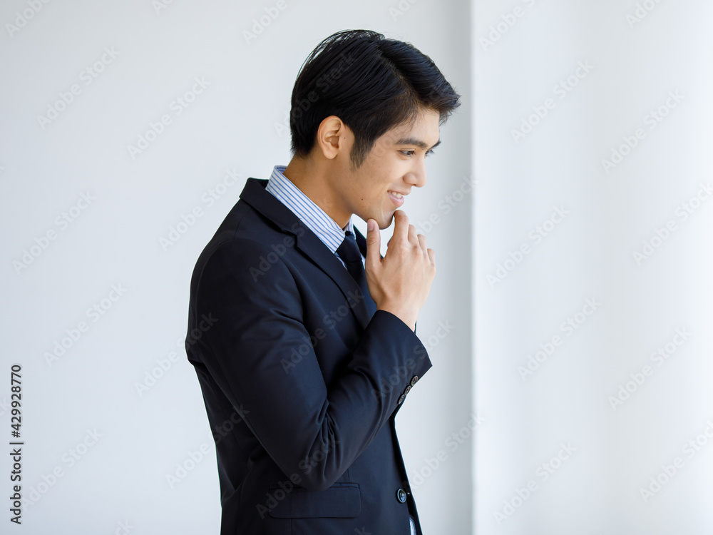 Portrait closeup shot of Asian young happy company leader in formal black suit and necktie standing hold hand touch chin look outside thinking finding solution idea in front of white wall background Portrait closeup shot of Asian young happy company leader in formal black suit and necktie standing hold hand touch chin look outside thinking finding solution idea in front of white wall background