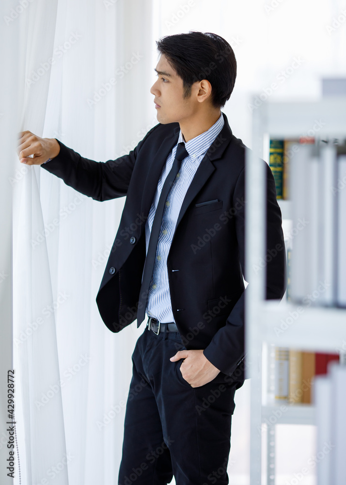 Portrait shot of Asian young company bookkeeper in formal black suit and necktie standing hold hand open clear white curtain look outside thinking brooding finding idea between bookshelf in library Portrait shot of Asian young company bookkeeper in formal black suit and necktie standing hold hand open clear white curtain look outside thinking brooding finding idea between bookshelf in library