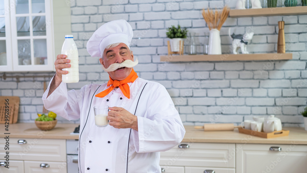 An elderly milkman holds a bottle of cow's milk in the kitchen. A man in a chef's hat is cooking
