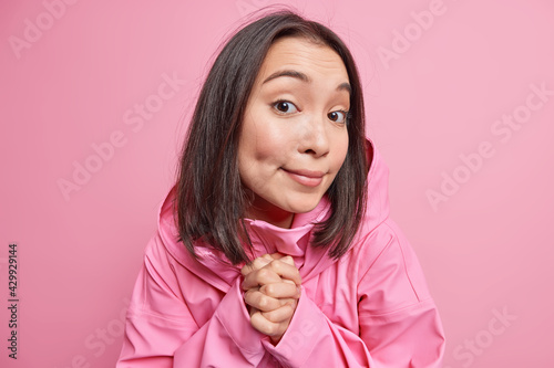 Photo of good looking young Asian woman with dark hair looks curiously at camera clasps hands together has curious gaze wears fashionable jacket isolated over pink background. Face expressions