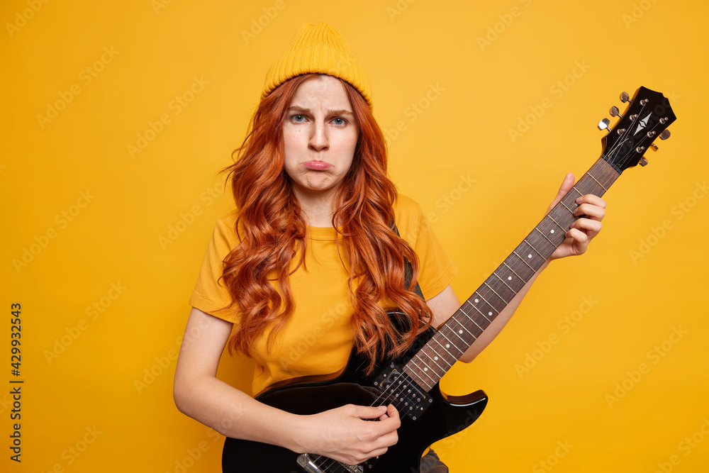 Dissatisfied offended redhead European woman purses lower lip plays electric guitar dressed casually poses with musical instrument isolated over yellow background. Music and singing concept. Dissatisfied offended redhead European woman purses lower lip plays electric guitar dressed casually poses with musical instrument isolated over yellow background. Music and singing concept.