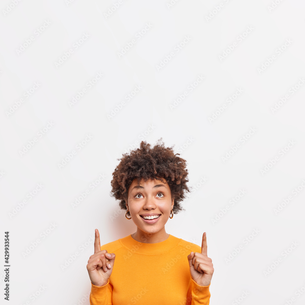 Photo of cheerful African American female points fingers up at top promo text suggests to check out awesome offer demonstrates copy space banner against white background wears casual jumper. - obrazy, fototapety, plakaty 