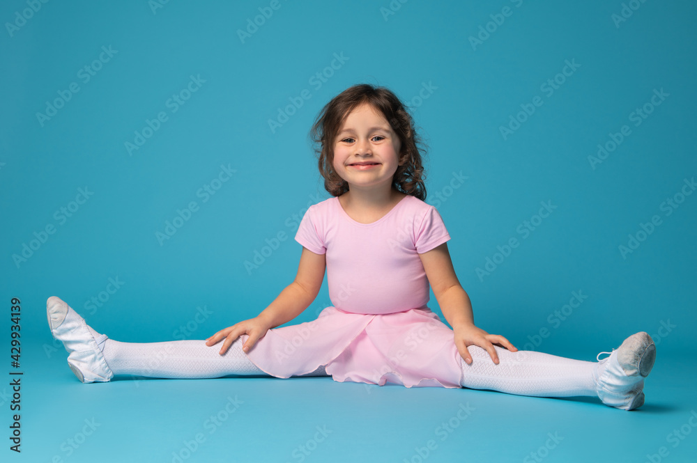 Beautiful little girl ballerina practices stretching her legs and cute smiles to the camera, isolated on blue background