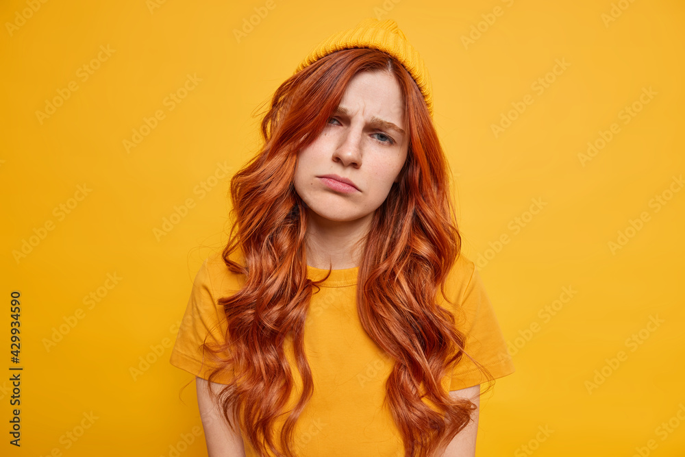 Frustrated sad redhead woman tilts head looks unhappily at camera purses lips feels indifferent wears hat and casual t shirt poses against vivid yellow background. Negative human emotions concept Frustrated sad redhead woman tilts head looks unhappily at camera purses lips feels indifferent wears hat and casual t shirt poses against vivid yellow background. Negative human emotions concept