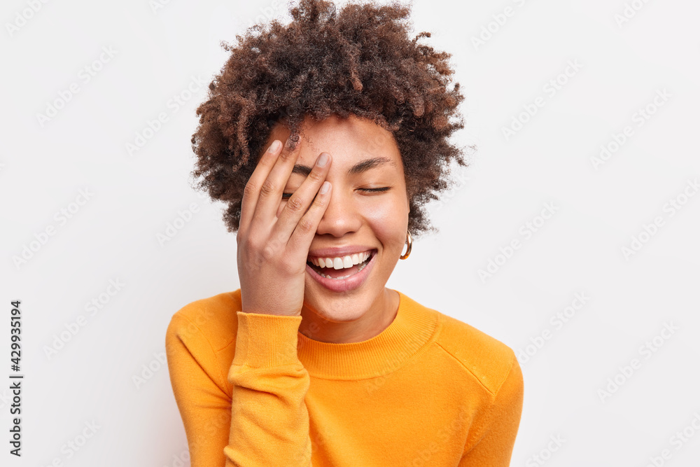 Close up shot of joyful carefree young woman with curly Afro hair smiles toothily keeps eyes closed makes face palm wears orange jumper expresses happiness isolated over white studio background Close up shot of joyful carefree young woman with curly Afro hair smiles toothily keeps eyes closed makes face palm wears orange jumper expresses happiness isolated over white studio background