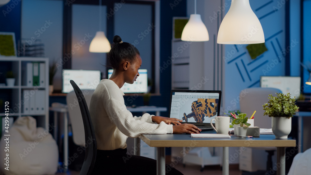 Fototapeta premium African engineer working late at night on a 3D model of industrial gears at laptop doing overtime in start up office. Freelancer studying prototype idea on pc showing cad software on device display