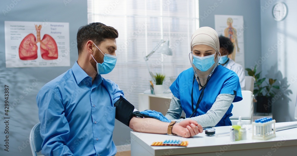 Pretty young Arab female nurse sitting in cabinet in hospital measuring blood tension to Caucasian man patient. Healthcare worker in medical center during covid-19 quarantine. Health concept Pretty young Arab female nurse sitting in cabinet in hospital measuring blood tension to Caucasian man patient. Healthcare worker in medical center during covid-19 quarantine. Health concept