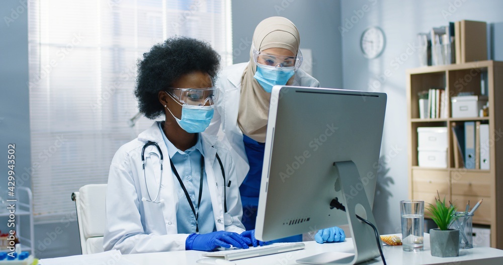 Portrait of mixed-race healthcare specialists working in cabinet in clinic. Arabic woman nurse speaking with African American young physician typing on computer browsing online. Hospital concept Portrait of mixed-race healthcare specialists working in cabinet in clinic. Arabic woman nurse speaking with African American young physician typing on computer browsing online. Hospital concept