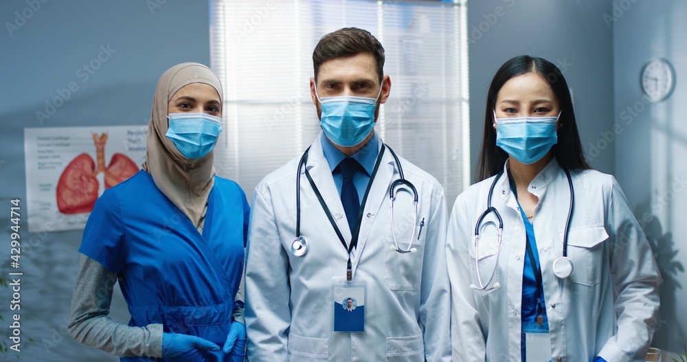Portrait of multi-ethnic cheerful young doctors in medical masks standing in medical center looking at camera and smiling. Team of physicians in clinic during coronavirus pandemic. Covid concept Portrait of multi-ethnic cheerful young doctors in medical masks standing in medical center looking at camera and smiling. Team of physicians in clinic during coronavirus pandemic. Covid concept
