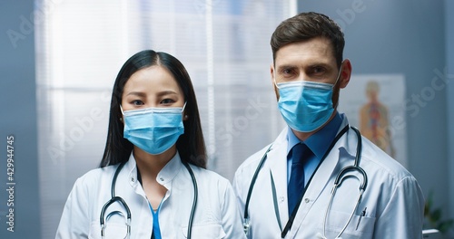Close up portrait of mixed-race couple of doctors wearing medical masks in coronavirus quarantine looking at camera and smiling in clinic. Caucasian man and pretty Asian woman physicians in hospital