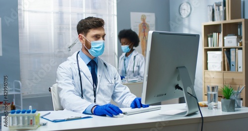 Portrait of Caucasian young handsome man professional physician at workplace in coronavirus pandemic sitting at table in cabinet texting on computer browsing online looking at monitor Hospital concept