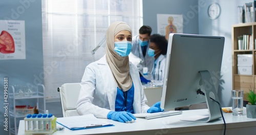 Young Arab beautiful professional female physician in medical mask working  typing and browsing online on computer sitting at workplace. Covid-19 virus  healthcare  medicine concept