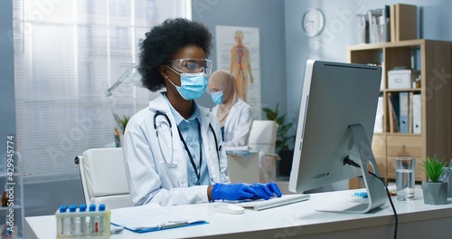 Young African American beautiful professional female physician in medical mask and goggles working, typing and browsing online on computer sitting at workplace. Covid-19 virus, medicine concept