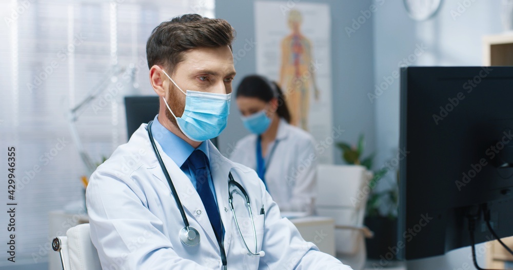 Close up portrait of young Caucasian handsome man doctor in medical mask working on computer sitting in hospital at workplace during quarantine looking at camera. Medical center, healthcare, covid-19 Close up portrait of young Caucasian handsome man doctor in medical mask working on computer sitting in hospital at workplace during quarantine looking at camera. Medical center, healthcare, covid-19