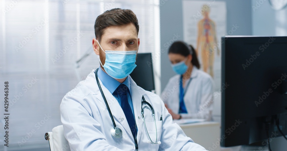 Close up portrait of young Caucasian handsome man doctor in medical mask working on computer sitting in hospital at workplace during quarantine looking at camera. Medical center, healthcare, covid-19 Close up portrait of young Caucasian handsome man doctor in medical mask working on computer sitting in hospital at workplace during quarantine looking at camera. Medical center, healthcare, covid-19
