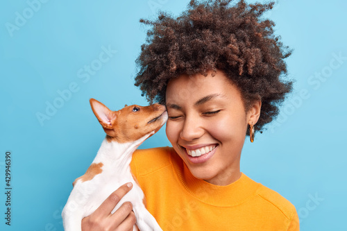 Friend of family. Close up shot of happy curly haired woman plays with dog expresses positive emotions likes animals. Small pedigree puppy licks face of owner. Adopted pet. Tender sincere feelings