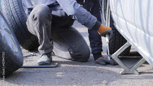 a man squatting on the asphalt lifts the car body with a screwdriver and a folding jack, wheel replacement and repair in road conditions, seasonal wheel change on his own