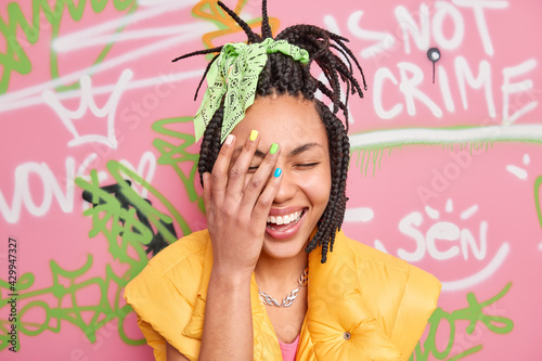 Cheerful milennial girl laughs sincerely makes face palm feels very happy has trendy hairstyle dressed in casual clothes poses against colorful graffiti wall. Happy female tagger at urban place