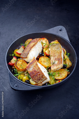 Modern style traditional fried skrei cod fish filet with fried potato, fruits and olives served as close-up in a design cast iron pan