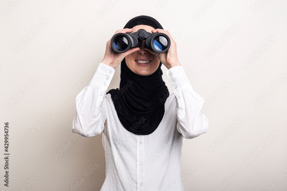 Friendly young Muslim woman in white shirt and hijab looks through binoculars. Banner. Looks into the camera Friendly young Muslim woman in white shirt and hijab looks through binoculars. Banner. Looks into the camera