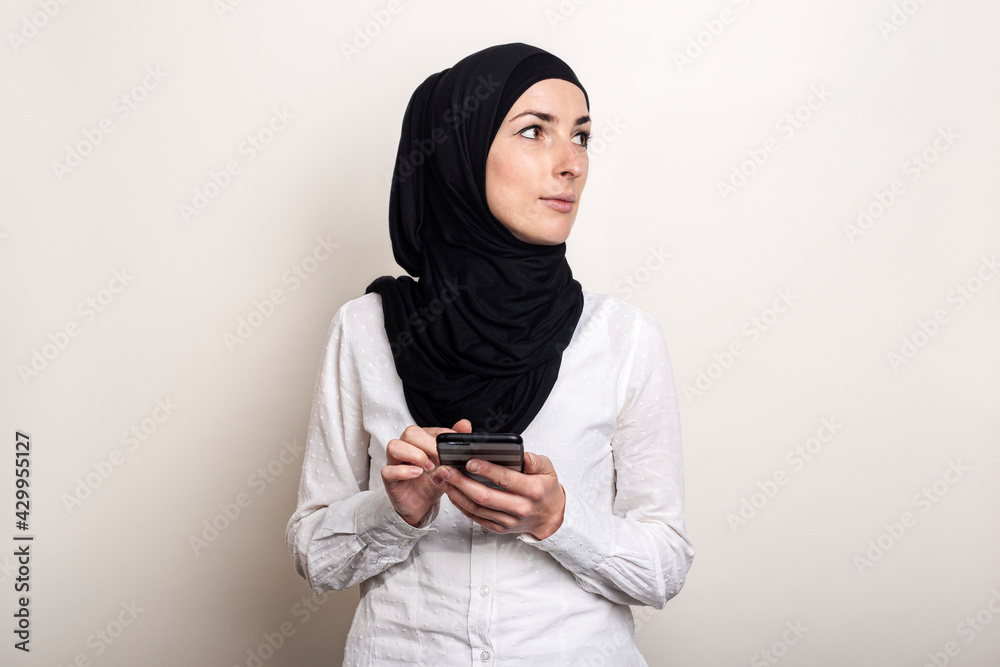 Muslim Young woman in hijab holds a phone in her hands and looks to the side on a light background. Banner