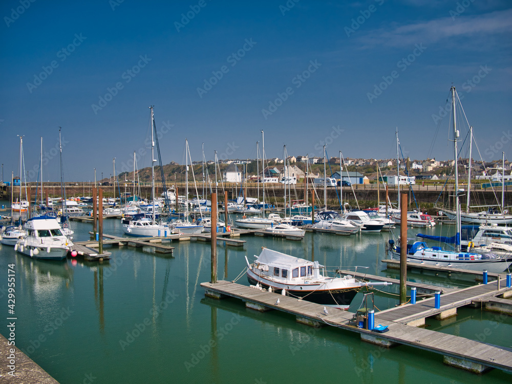 In sunshine on a calm sunny day, leisure boats moored on piers at Maryport Marina in north west Cumbria, England, UK. In the calm water are reflections of the boats and their masts.