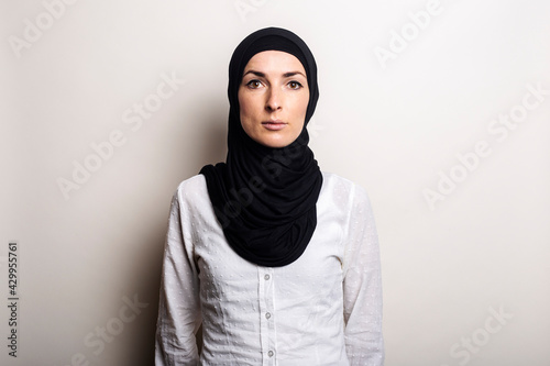 Young woman in a white shirt and hijab on a light background. Looks into the camera