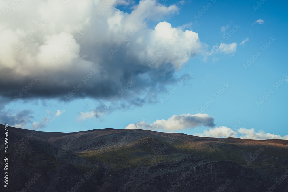 Minimalist alpine landscape with mountain silhouette under cloudy sky with big cloud. Dark mountain and light sky. Minimal nature background with silhouette of mountain under large cloud in blue sky.