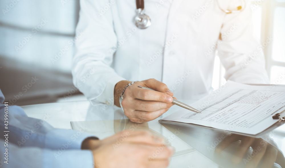 Unknown male doctor and woman-patient discussing current health examination while sitting in sunny clinic and using clipboard. Good medical service in hospital Unknown male doctor and woman-patient discussing current health examination while sitting in sunny clinic and using clipboard. Good medical service in hospital