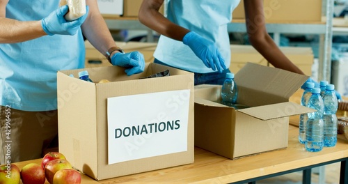 Close up of Caucasian male and African American female hands in gloves packing food and grocery in boxes preparing donation delivery. Charity activity, social help for poor, volunteering and donating © VAKSMANV
