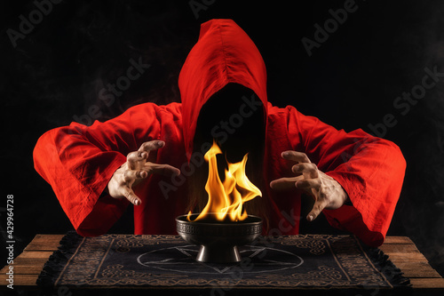 Long haired male making a mystical flames. Man dressed in a red on a dark smoky background. Witch or wizard with fire in hands. Mysticism and black magic concept.  photo