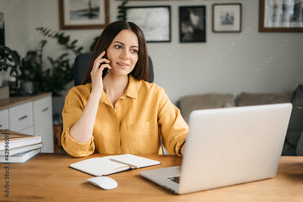 Smiling business woman talking on a mobile phone and using a laptop while working at home, looking at the screen, reading a message, a young female employee advises a client, telephone conversations Smiling business woman talking on a mobile phone and using a laptop while working at home, looking at the screen, reading a message, a young female employee advises a client, telephone conversations