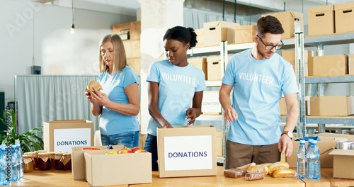 Slika na platnu Busy African American and Caucasian female and male volunteers preparing donations box, packing food and grocery