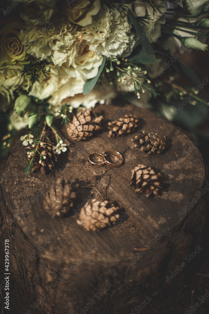 Gold wedding rings on a round wooden stump in the forest, surrounded by fir cones. Nearby, a beautiful bouquet of white and beige roses lies on the ground. Brown, white, beige. Vertical close frame. Gold wedding rings on a round wooden stump in the forest, surrounded by fir cones. Nearby, a beautiful bouquet of white and beige roses lies on the ground. Brown, white, beige. Vertical close frame.