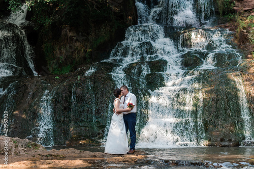 The bride and groom stand against the background of a waterfall. Newlyweds on the background of a waterfall