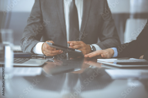 Unknown businessman using tablet computer and working together with his colleague while sits at the glass desk in modern office. Teamwork and partnership concept