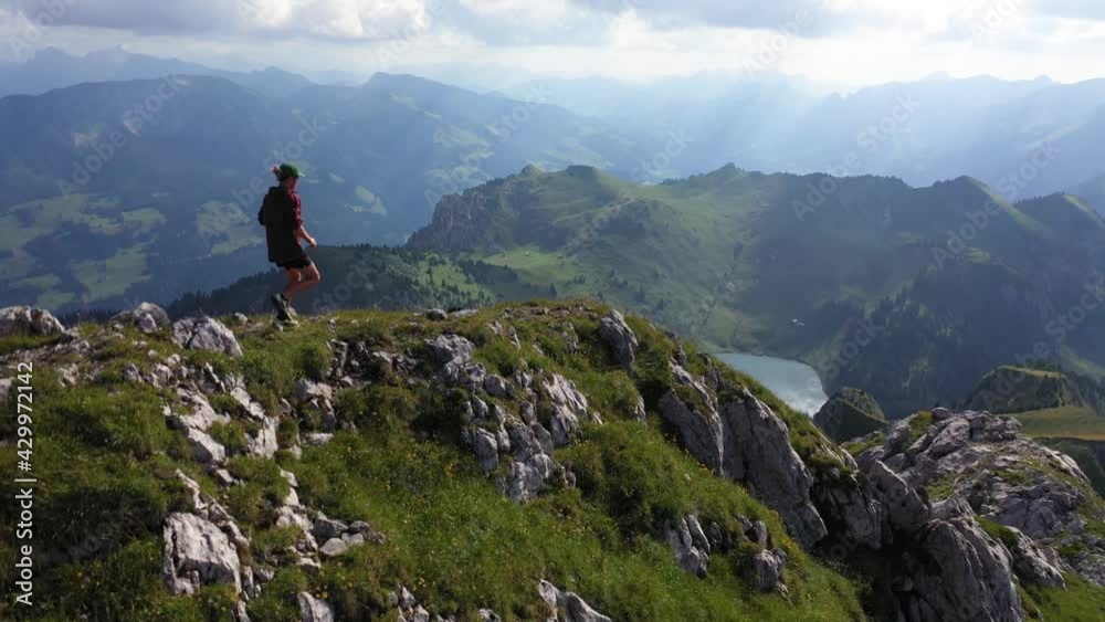 Drone flying backwards while a young, fit man is running down the rocky trail on the edge of the ridge. Insignificant and small compared to the majestic and vast landscape in the hot sunny weather.