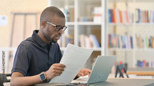 Young African Man with Laptop doing Paperwork in Library